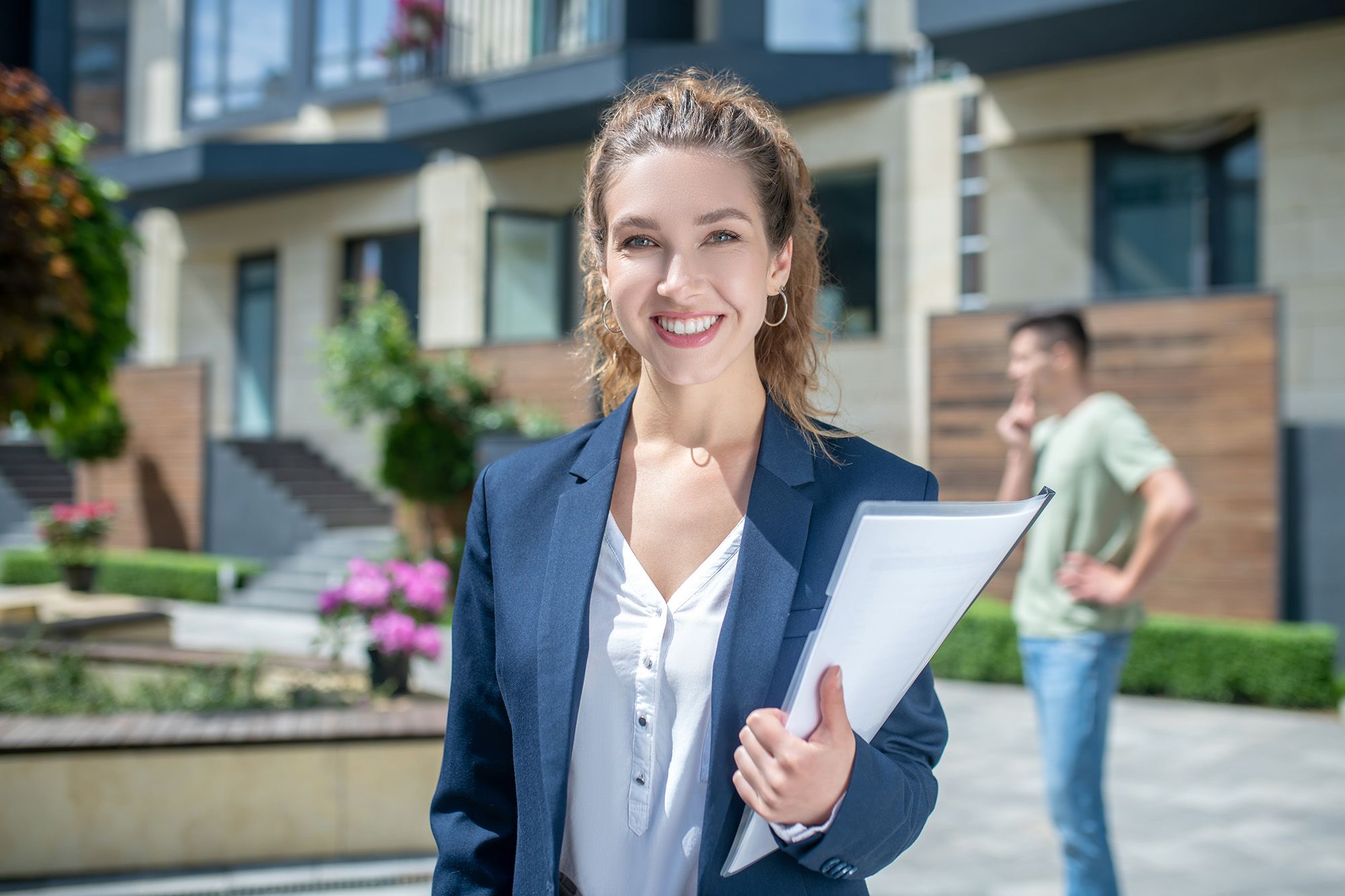 Smiling professional real estate agent holding documents in front of a modern property, symbolizing career growth and client-focused service